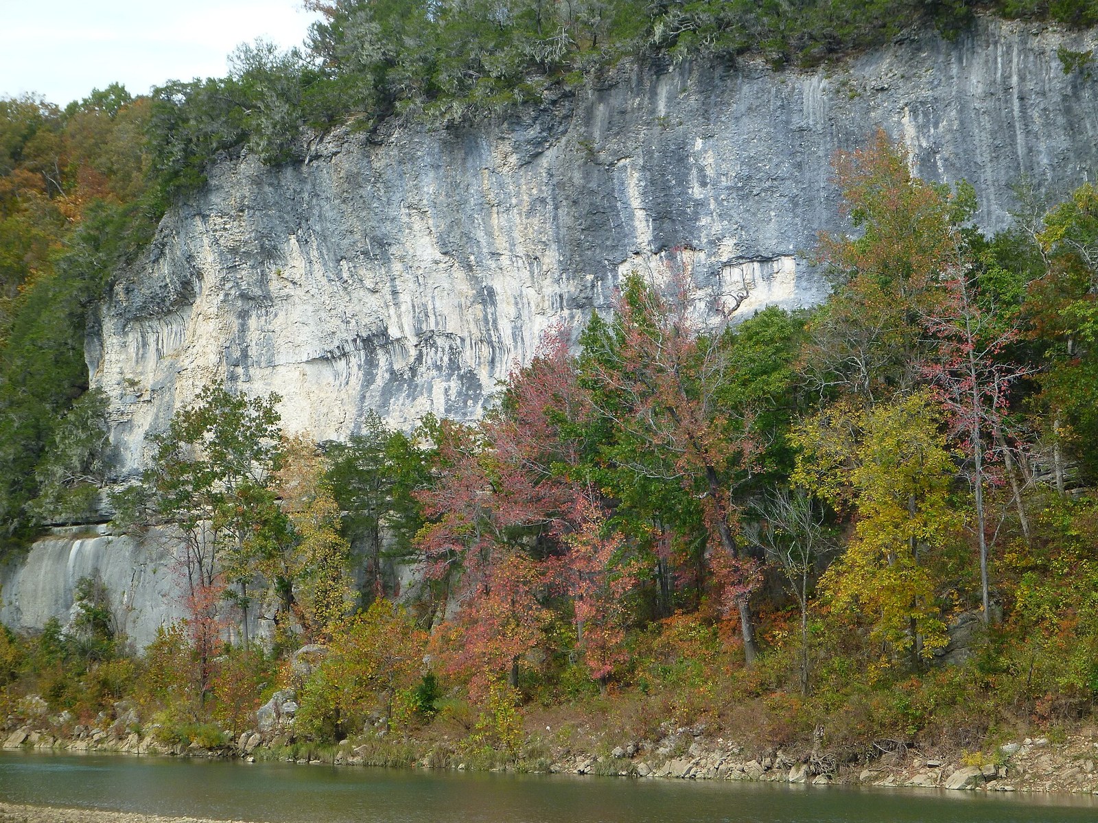 A tall gray bluff rising over green water, with red, orange and yellow trees along its base.