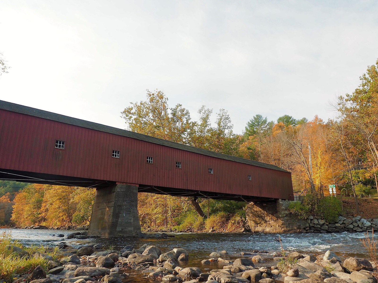 A long red covered bridge over a rocky river, with orange and yellow trees along the far bank.