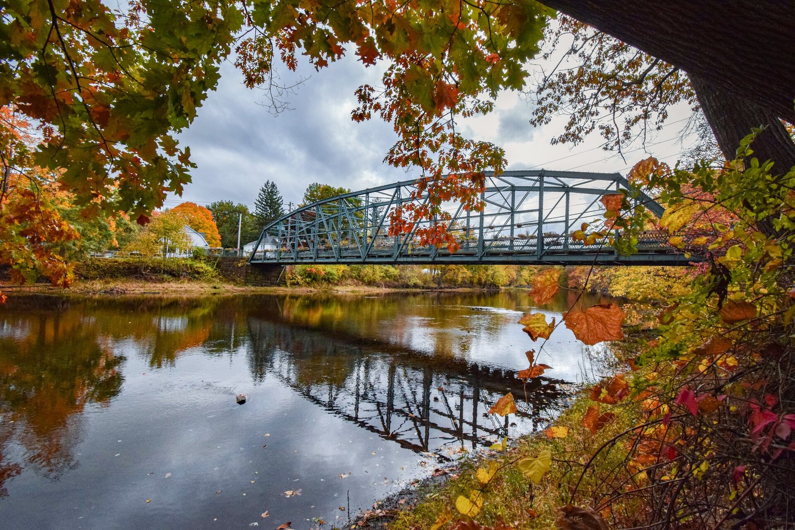 A steel truss bridge over a still river, framed by red, orange and yellow maple leaves.