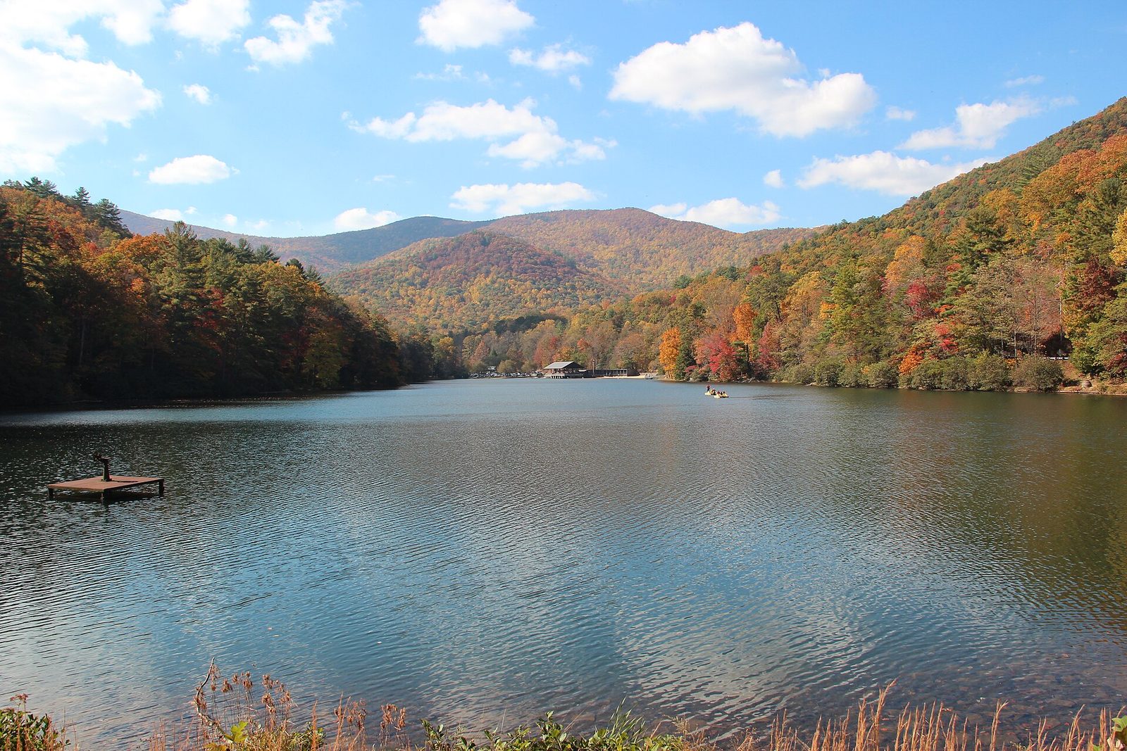 A calm lake in a mountain valley, with hillsides of orange, red and gold trees under a blue sky.
