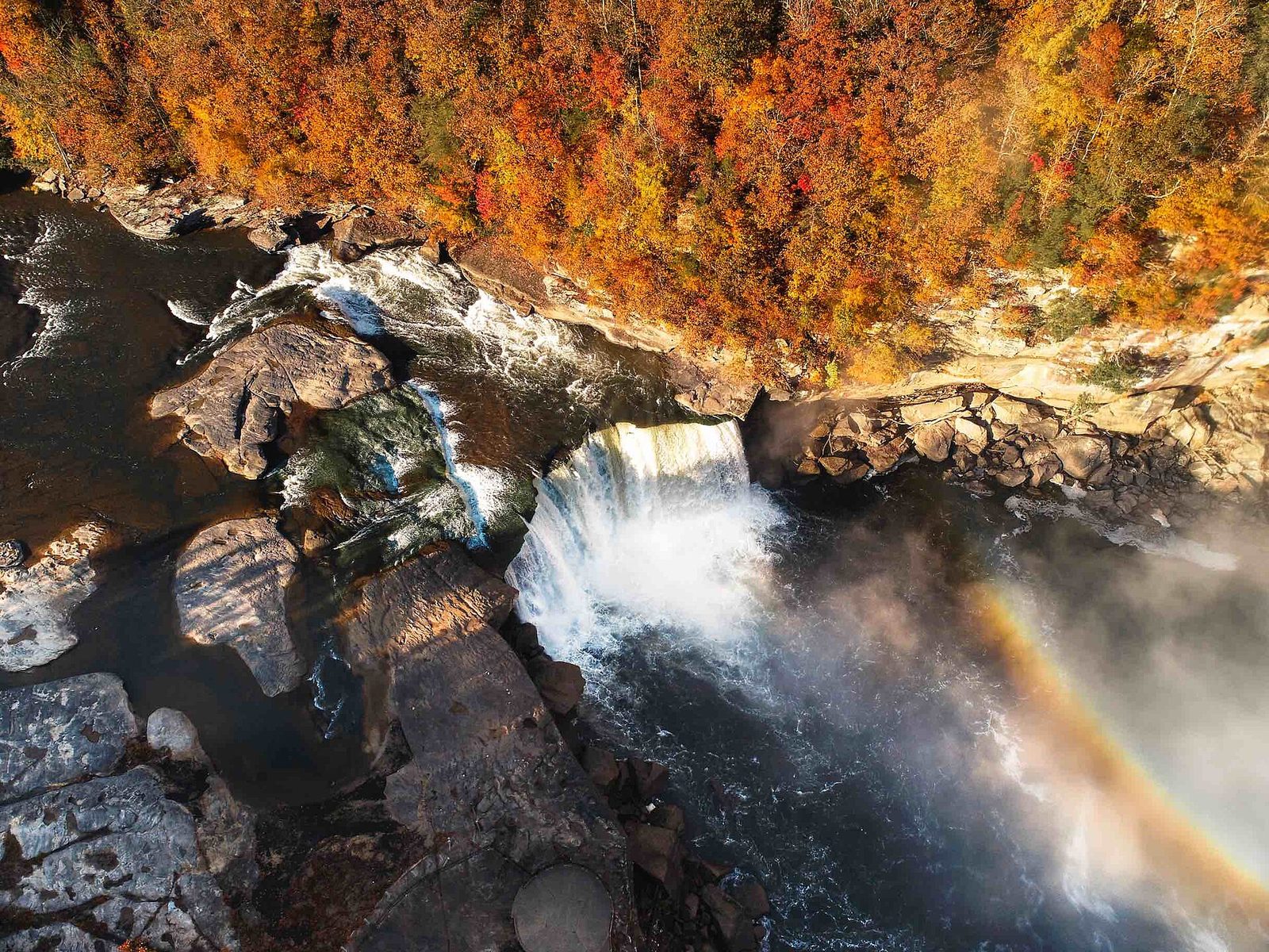 Cumberland Falls seen from above, with a rainbow in the mist and orange and gold trees along the cliff.