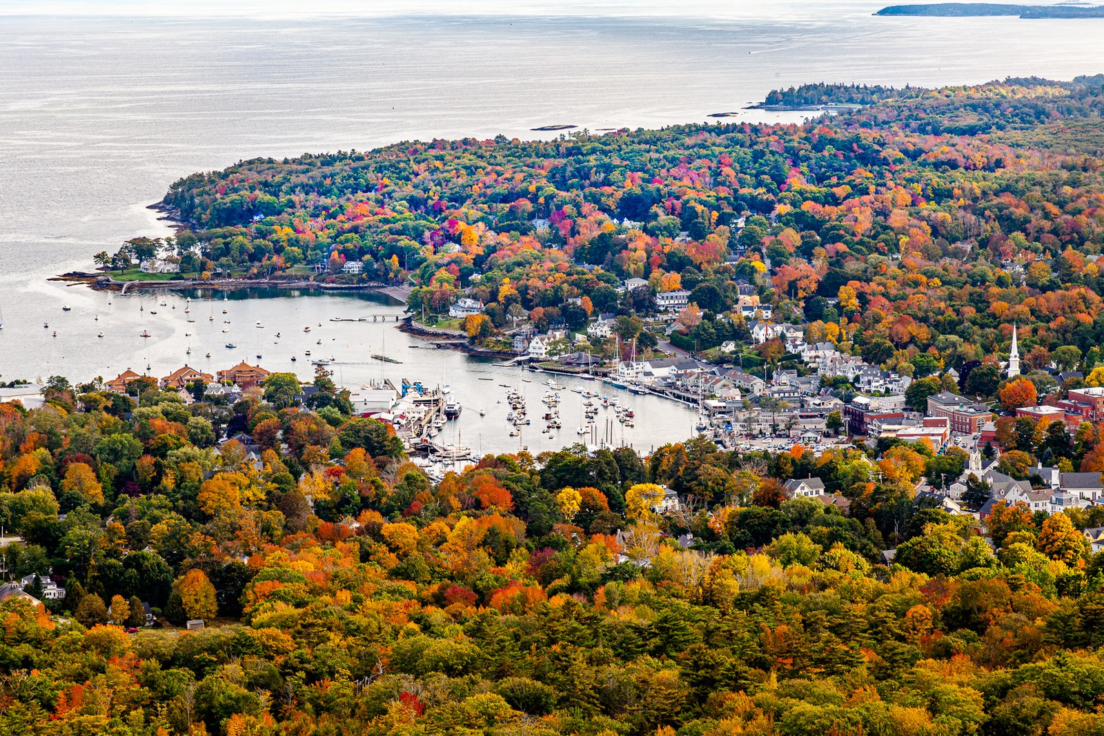 Camden harbor and the town among red, orange and gold trees, seen from above, with the bay beyond.