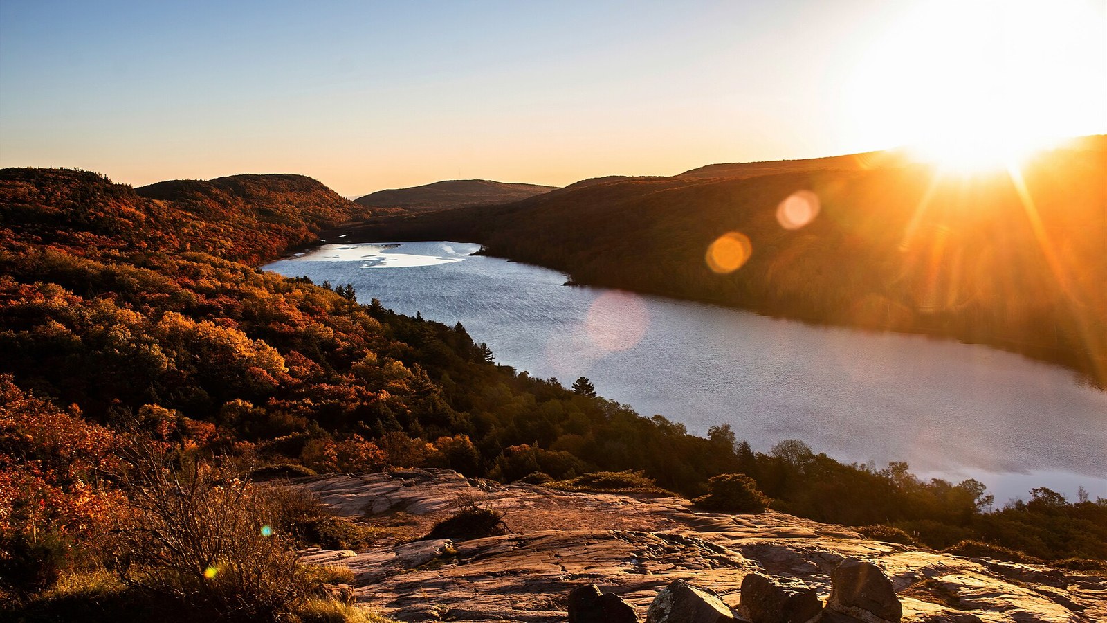 Sunset over a long lake in a forested valley, with red and orange hillsides in the foreground.