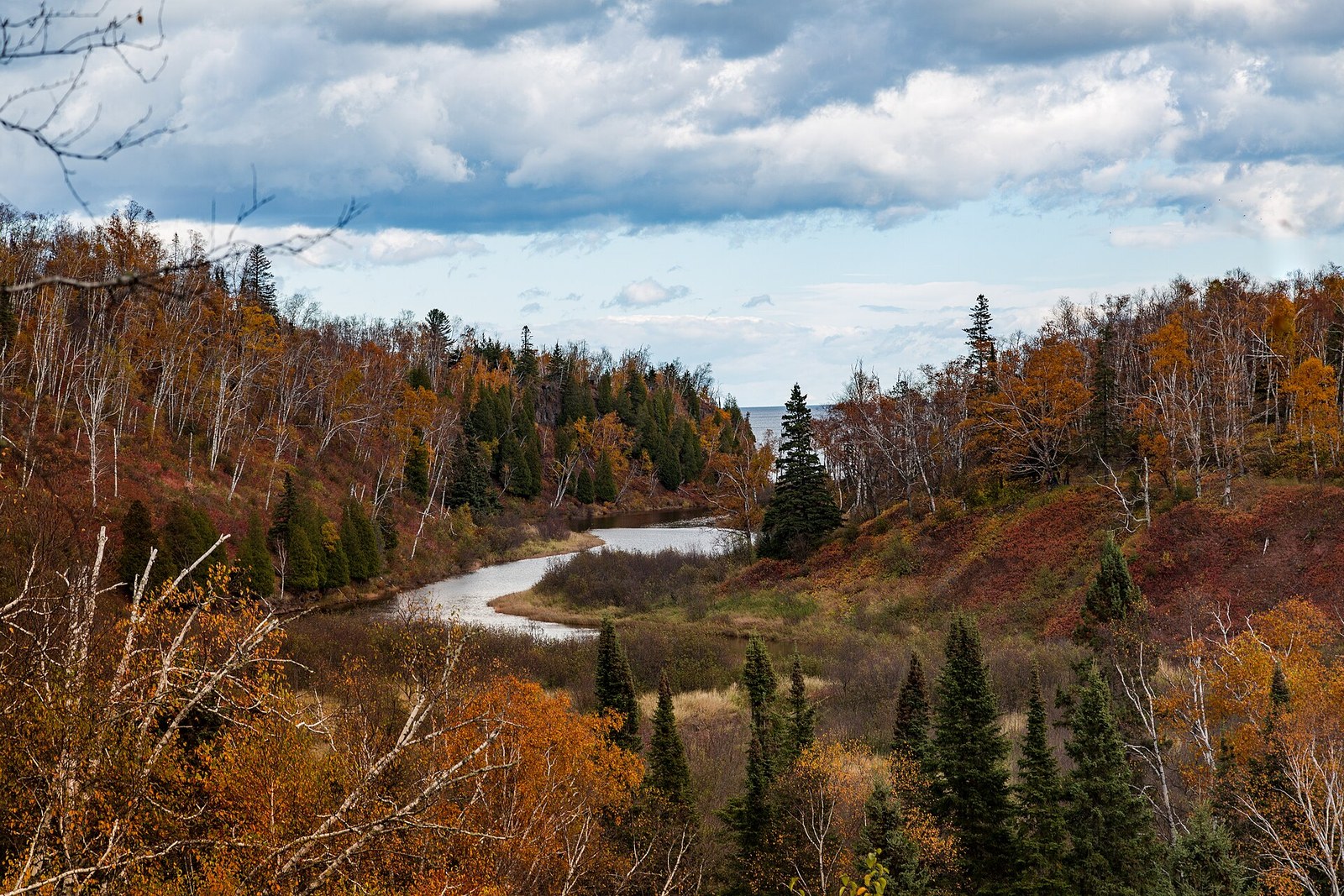 A river bending through a hillside of orange and red birch and aspen with dark spruce, and a lake horizon at the far right.