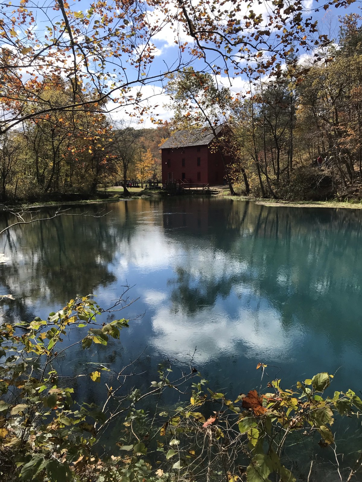 A red mill beside a clear blue-green spring, with orange and yellow leaves on the trees around it.