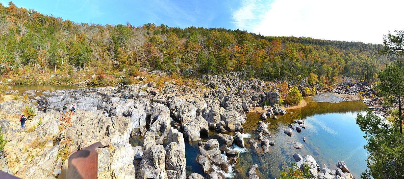 Pink-gray rocks of the shut-ins in a river, with hillsides of gold, orange and green trees behind.