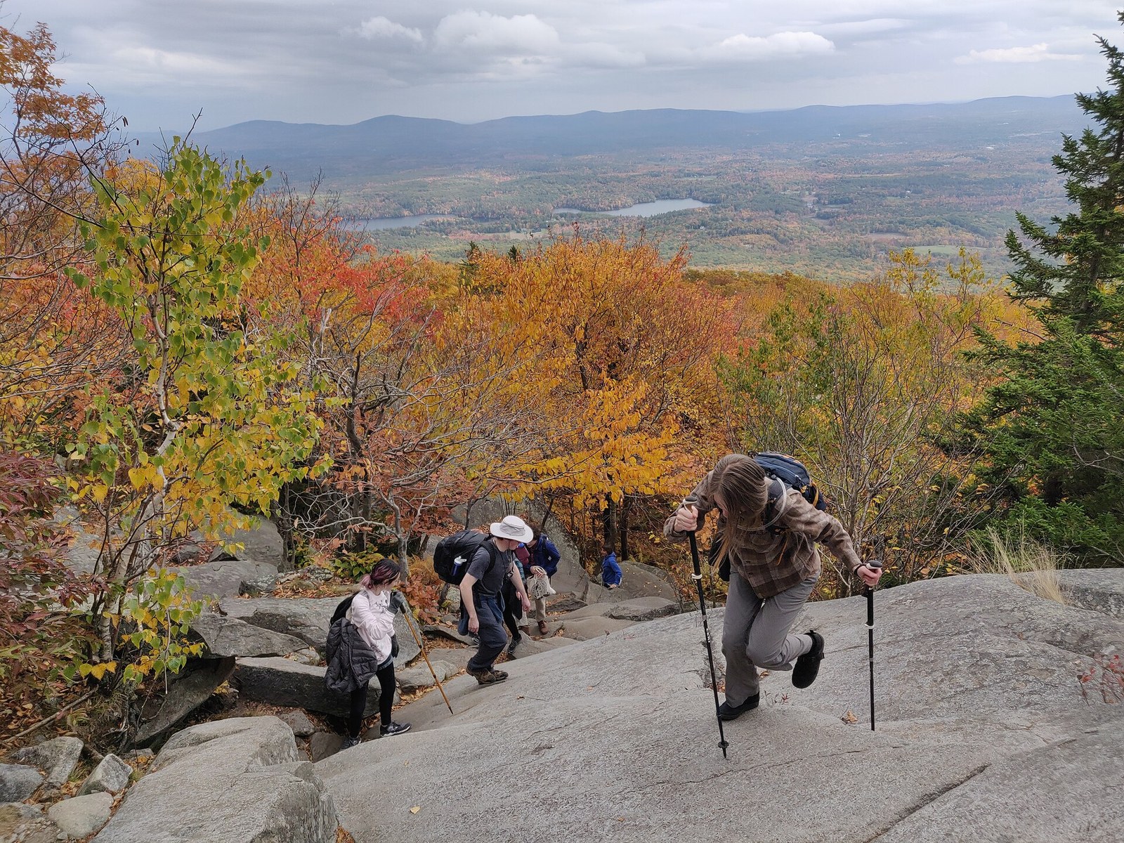 Hikers climbing a granite slope past trees in gold, orange and red, with a wide valley below.