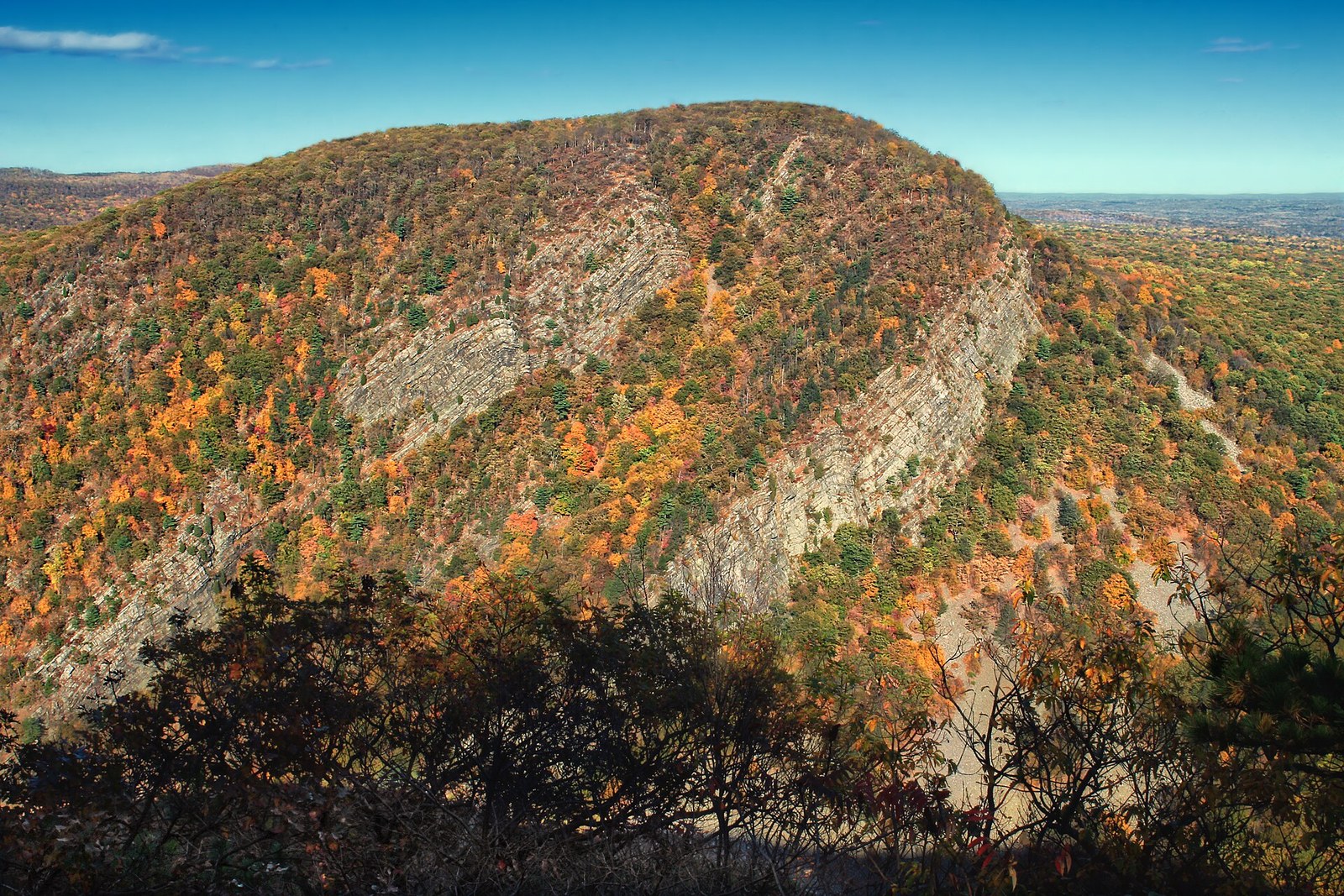 A steep, rocky mountain covered in orange, yellow and green trees, seen across a gap.